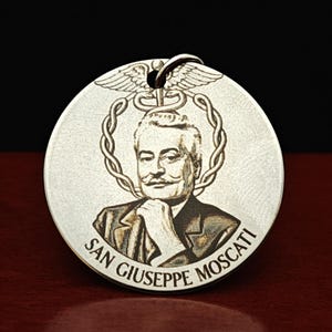 Op de afbeelding: Een ronde, zilverkleurige metalen hanger met een gegraveerd portret van San Giuseppe Moscati. Boven het portret staat een caduceussymbool met vleugels en slangen. De tekst SAN GIUSEPPE MOSCATI is onderaan de rand van de hanger gegraveerd.