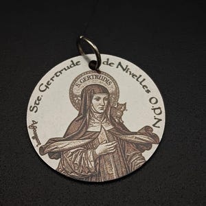 May include: Circular pendant depicting St. Gertrude of Nivelles, with a halo and an owl. The pendant has a metal ring for hanging and text around the image. The image is in sepia tones.
