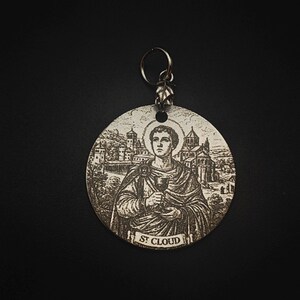 Op de afbeelding: Een ronde metalen hanger met een gegraveerde afbeelding van een figuur die een kelk vasthoudt, met een aureool boven het hoofd en een gebouw op de achtergrond. De tekst ST CLOUD staat op een banier aan de onderkant. Een metalen ring is bevestigd.