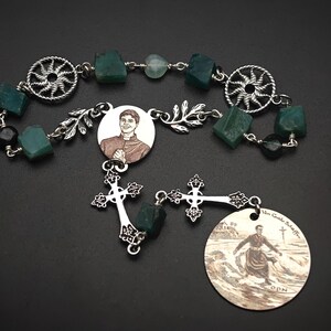 May include: A rosary featuring green gemstone beads, silver-toned metal accents, and a central medallion with a portrait. The design includes decorative crosses and a circular pendant with an image of a person.