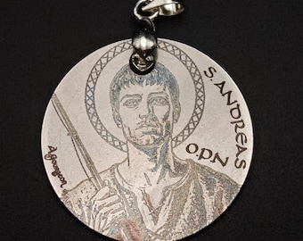 May include: A silver pendant featuring an engraved image of Saint Andrew, with the text "S. Andreas O.P.N." around the edge. The pendant has a bail for attaching to a chain.