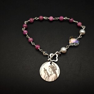 May include: A silver-toned bracelet featuring a round metal charm engraved with the image of St. Mary Magdalene and the text St. Mary Magdalene OPN. The chain includes faceted magenta beads, two white pearls, and a clear iridescent crystal bead.