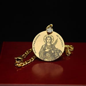 May include: A gold-toned circular pendant featuring an engraved image of Saint Margaret of Antioch, with the text "SAINT MARGARET OF ANTIOCH" inscribed around the edge. The pendant includes a small cross-shaped bail with embedded clear stones.
