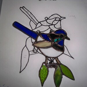 May include: A stained glass artwork depicting two blue wrens, one perched on top of the other. The birds are surrounded by green leaves and have a white background.