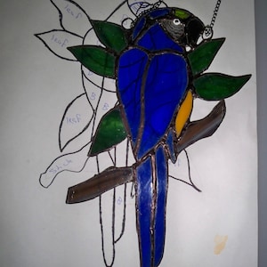 May include: A stained glass blue and yellow macaw perched on a branch with green leaves. The macaw is hanging from a chain.