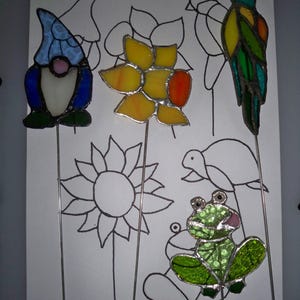 May include: Five stained glass pot plant stakes. The stakes feature a gnome, a flower, a sunflower, a frog, and a bird. The stakes are all on a white background with the words "Pot plant stakes" written at the top.