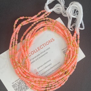 May include: Coral-colored beaded waist beads with gold accents and white string ties. The beads are arranged in multiple strands, resting on a white card with the text "COLLECTIONS" and "Handmade African waist beads and accessories".