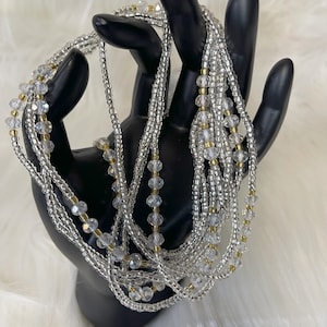 May include: A collection of silver and clear beaded waist beads with gold accents. The beads are draped over a black hand-shaped display, creating a layered effect. The beads are on a white fluffy surface.