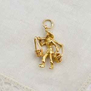 May include: Gold charm depicting a person carrying two buckets. The charm is detailed and has a vintage look.
