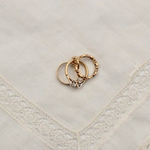 May include: Three gold rings stacked on top of each other on a white lace background. The top ring is a textured band, the middle ring is a plain band, and the bottom ring is a textured band with a small design.