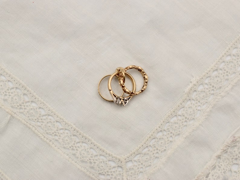 May include: Three gold rings stacked on top of each other on a white lace background. The top ring is a textured band, the middle ring is a plain band, and the bottom ring is a textured band with a small design.
