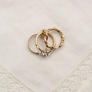May include: Three gold rings stacked on top of each other. The top ring has a textured design, the middle ring is plain, and the bottom ring has a decorative design with small stones.