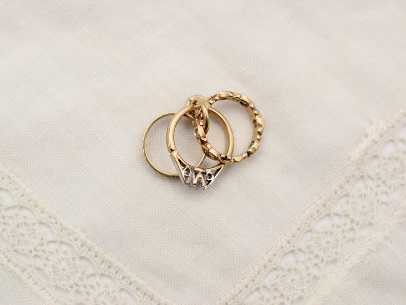 May include: Three gold rings stacked on top of each other. The top ring has a textured design, the middle ring is plain, and the bottom ring has a decorative design with small stones.