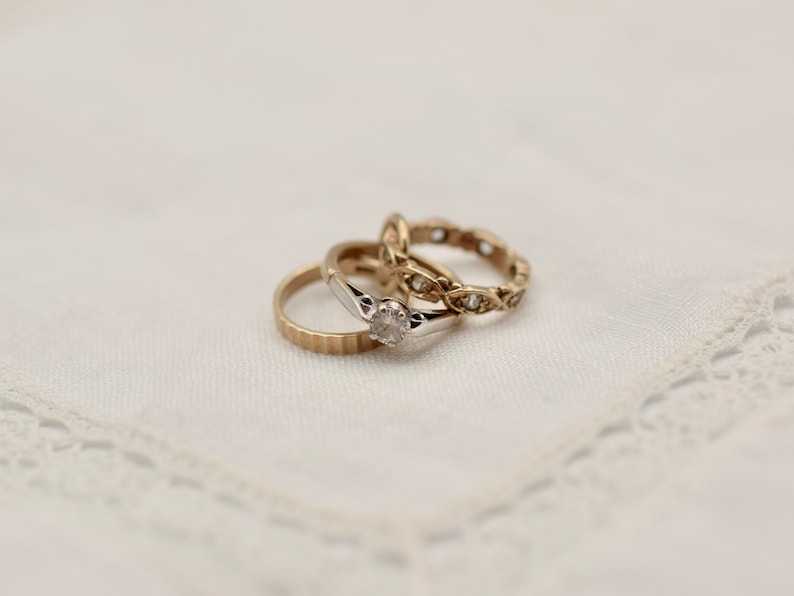 May include: Two gold rings with a diamond on one ring. The rings are on a white lace background.