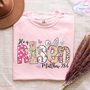 A pink t-shirt with the text "He is Risen Matthew 28:6" printed on it, along with various Easter-themed graphics such as bunny ears, butterflies, and flowers. The t-shirt is placed on a bed with a brown hat and a bunch of purple flowers nearby.