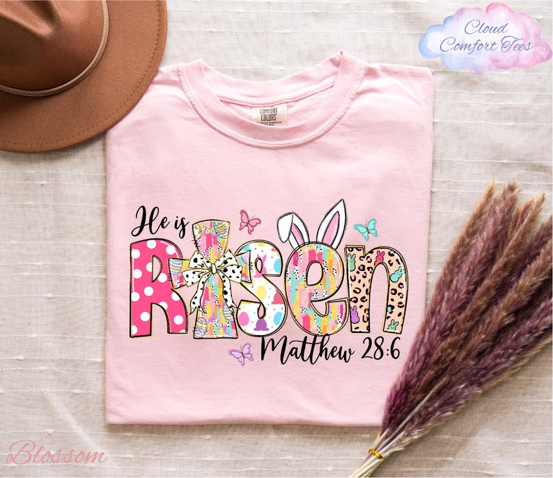 A pink t-shirt with the text "He is Risen Matthew 28:6" printed on it, along with various Easter-themed graphics such as bunny ears, butterflies, and flowers. The t-shirt is placed on a bed with a brown hat and a bunch of purple flowers nearby.