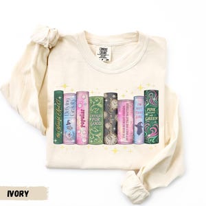 A white long-sleeved t-shirt with a graphic design of a row of colorful books with various titles and illustrations.
