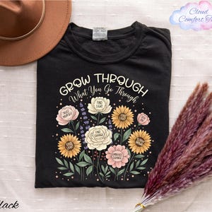 a black t-shirt with a floral design and the text "Grow Through What You Go Through" printed on it, along with a brown hat and some pom poms.