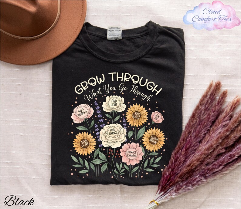 a black t-shirt with a floral design and the text "Grow Through What You Go Through" printed on it, along with a brown hat and some pom poms.
