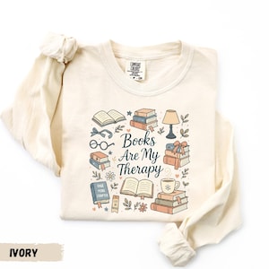 May include: Ivory-colored long-sleeve shirt with a graphic that reads "Books Are My Therapy." The design includes illustrations of books, glasses, a lamp, and a coffee cup. The shirt is made of a soft material.
