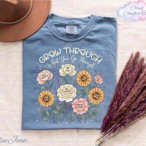 A blue t-shirt with a floral design and the text "Grow Through What You Go Through" printed on it, along with a brown hat and a bunch of purple flowers.