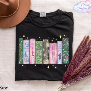 A black t-shirt with a graphic design of colorful books and a brown hat, along with some purple and green decorative elements, and a bunch of purple and green decorative elements.