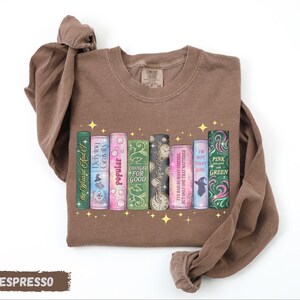 A brown t-shirt with a graphic design of colorful book spines arranged in a row, with the titles "Popular for good", "I've had so many friends that I'm not that girl", "I'm not pink that girl", "I'm not green that girl", and "I'm not that girl that girl".