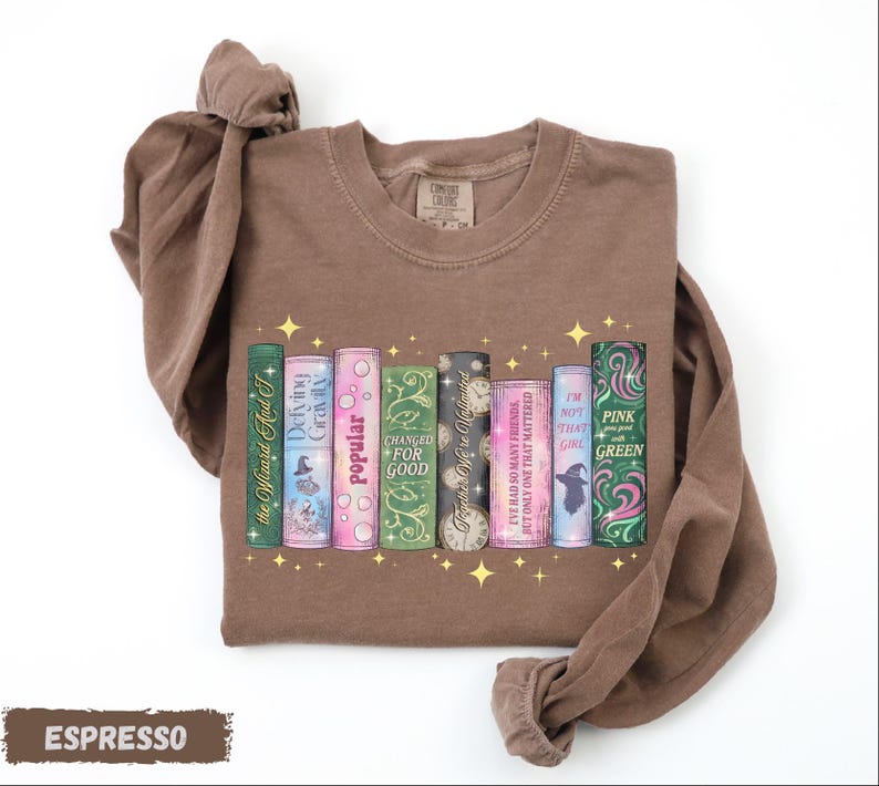 A brown t-shirt with a graphic design of colorful book spines arranged in a row, with the titles "Popular for good", "I've had so many friends that I'm not that girl", "I'm not pink that girl", "I'm not green that girl", and "I'm not that girl that girl".