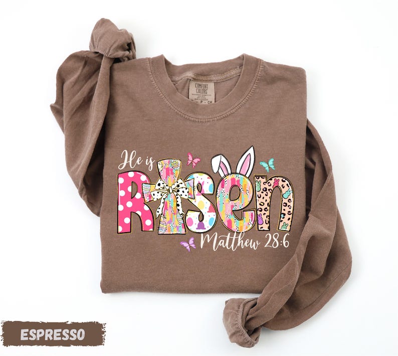 A brown t-shirt with a colorful Easter-themed design, including the text "He is Risen" and "Matthew 28:6".