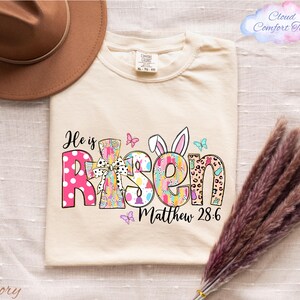 a beige t-shirt with a colorful Easter-themed design, including the text "He is Risen" and "Matthew 28:6". The t-shirt is laid out on a white surface, with a brown hat and some purple flowers nearby.