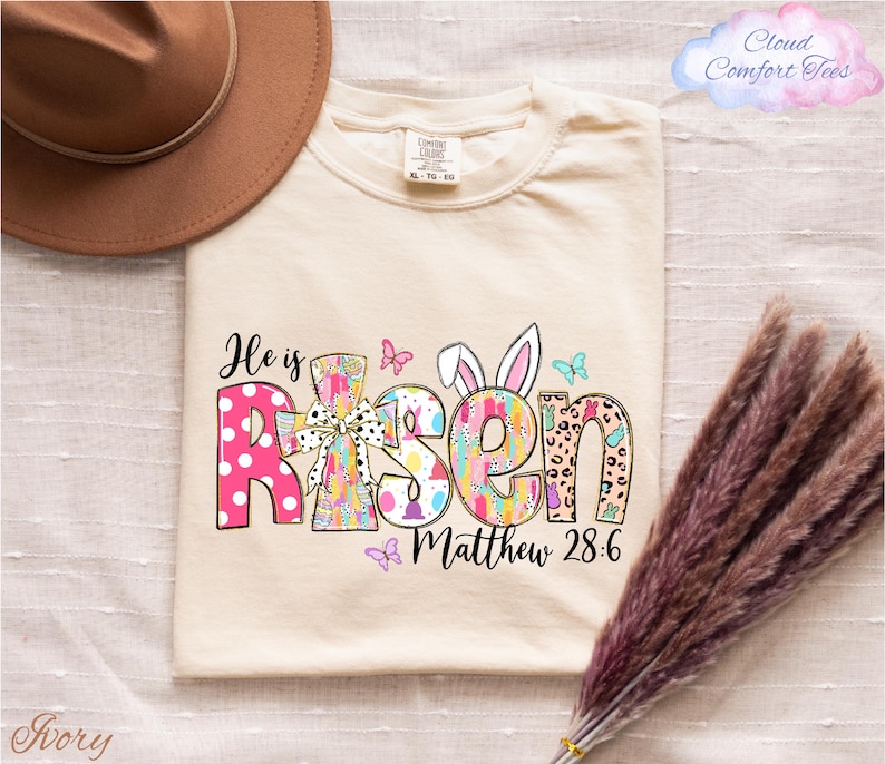 a beige t-shirt with a colorful Easter-themed design, including the text "He is Risen" and "Matthew 28:6". The t-shirt is laid out on a white surface, with a brown hat and some purple flowers nearby.