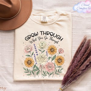 a white t-shirt with a floral design and the text "Grow Through What You Go Through" printed on it, along with a brown hat and some dried flowers.
