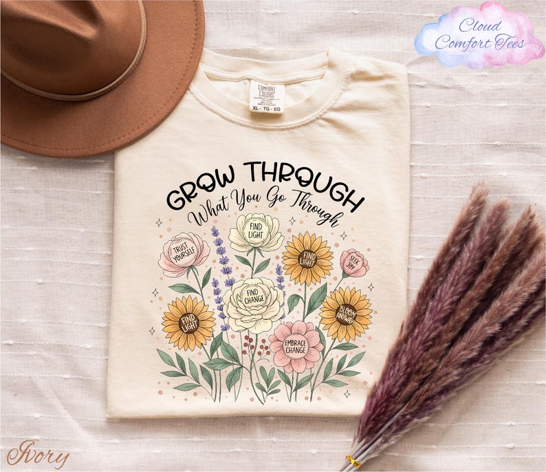 a white t-shirt with a floral design and the text "Grow Through What You Go Through" printed on it, along with a brown hat and some dried flowers.