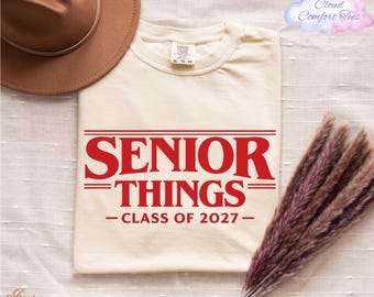May include: An ivory-coloured t-shirt with the words "SENIOR THINGS" in red, and "CLASS OF 2027" below. A brown hat and dried flowers are in the background. The shirt is a Comfort Colors brand.