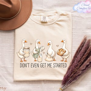 May include: An ivory t-shirt featuring a graphic of four white geese engaged in baking activities, including holding a sourdough jar, mixing in a bowl, and carrying a loaf of bread. The text below the geese reads, DON'T EVEN GET ME STARTED.