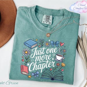 May include: Light green t-shirt with the text "Just one more chapter" surrounded by illustrations of books, flowers, and a coffee cup. The shirt has a crew neck and short sleeves. A gold necklace is draped across the shirt.