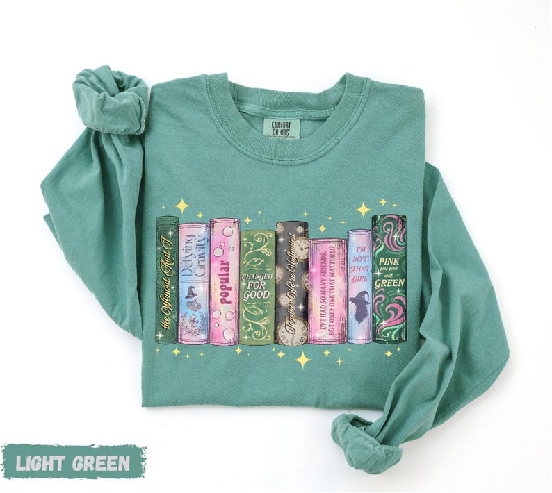 A light green long-sleeved t-shirt with a graphic design of a row of colorful books with various titles and illustrations.