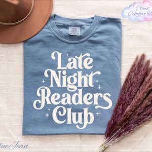 May include: A blue jean colored t-shirt with the text "Late Night Readers Club" in white script. The shirt is folded and has a brown hat and dried flowers in the image.