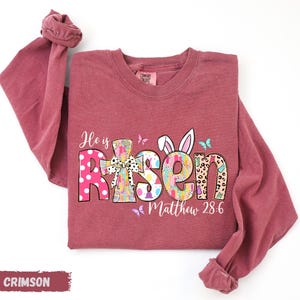 A red long-sleeved t-shirt with a colorful Easter-themed design, including the text "He is Risen" and "Matthew 28:6".