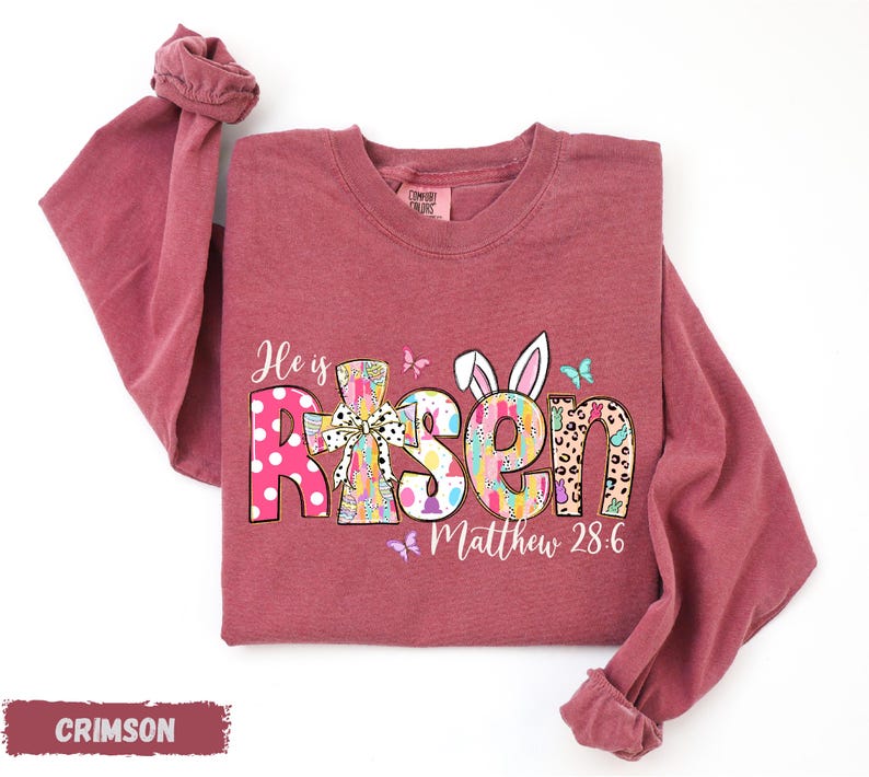 A red long-sleeved t-shirt with a colorful Easter-themed design, including the text "He is Risen" and "Matthew 28:6".