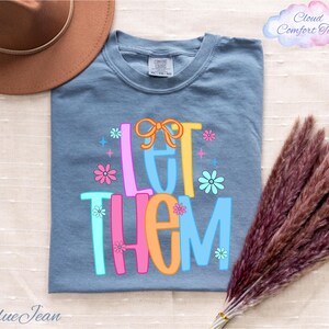 May include: Blue jean colored t-shirt with the words "LET THEM" in colorful, playful lettering. The design includes flowers and a bow. The shirt is part of the Cloud Comfort Tees collection.
