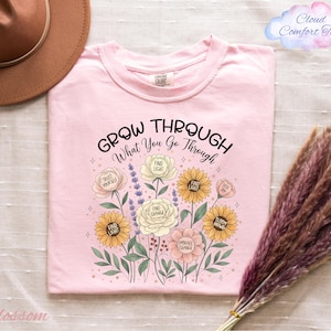 a pink t-shirt with a floral design and the text "Grow Through What You Go Through" printed on it, along with illustrations of various flowers and leaves. The t-shirt is placed on a bed with a brown hat and a bunch of purple flowers nearby.