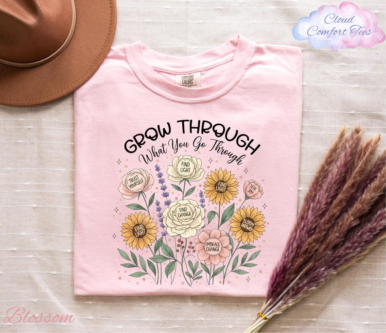 a pink t-shirt with a floral design and the text "Grow Through What You Go Through" printed on it, along with illustrations of various flowers and leaves. The t-shirt is placed on a bed with a brown hat and a bunch of purple flowers nearby.
