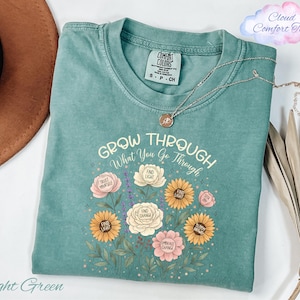 a light green t-shirt with a floral design and the text "Grow Through" printed on it. The t-shirt is placed on a white surface, along with a brown hat, a necklace, and some dried leaves.
