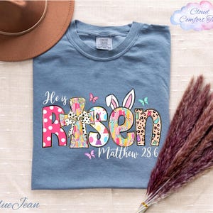 a blue t-shirt with a colorful Easter-themed design, including the text "He is Risen" and "Matthew 28:6". The t-shirt is laid out on a surface, along with a brown hat and some purple reeds.
