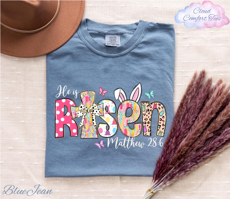 a blue t-shirt with a colorful Easter-themed design, including the text "He is Risen" and "Matthew 28:6". The t-shirt is laid out on a surface, along with a brown hat and some purple reeds.