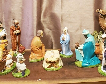May include: A nativity scene set with figurines of Mary, Joseph, baby Jesus, three wise men, shepherds, and animals. The set includes a camel, a donkey, and sheep. The figurines are made of painted ceramic and are arranged on a wooden surface.