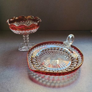 May include: Two clear glass serving dishes with a red, iridescent finish. The larger dish has a scalloped edge and a pedestal base. The smaller dish has a handle and a scalloped edge.