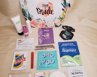 May include: A white toiletry bag with the words "Team Bride" in pink lettering. The bag is surrounded by various items, including makeup remover wipes, cotton swabs, nail polish remover pads, bandages, and a small bottle of hand sanitizer.