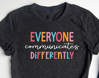 May include: A dark gray t-shirt with the text "EVERYONE communicates DIFFERENTLY" in colorful letters. The shirt is laid flat on a white surface with a pair of jeans, a brown purse, a camera, and a potted succulent.
